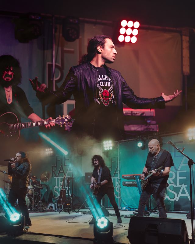 Rock band on stage under teal beams, the singer opening his arms to the crowd