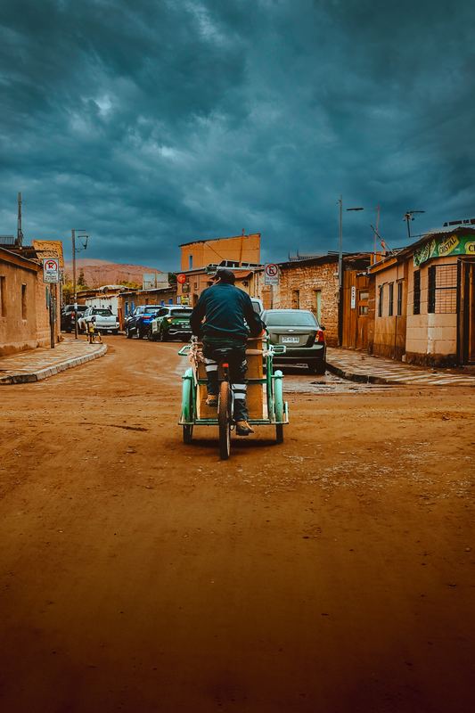 A dirt street under a storm sky, a man riding away on a three-wheeler