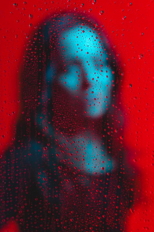 Volto di donna dietro un vetro coperto di gocce di pioggia, rosso e ciano